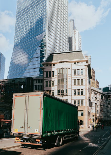 A large green and beige moving van positioned on a city street in Shoreditch with modern glass skyscrapers and older stone buildings in the background. The van is parked near a curb, indicating a load